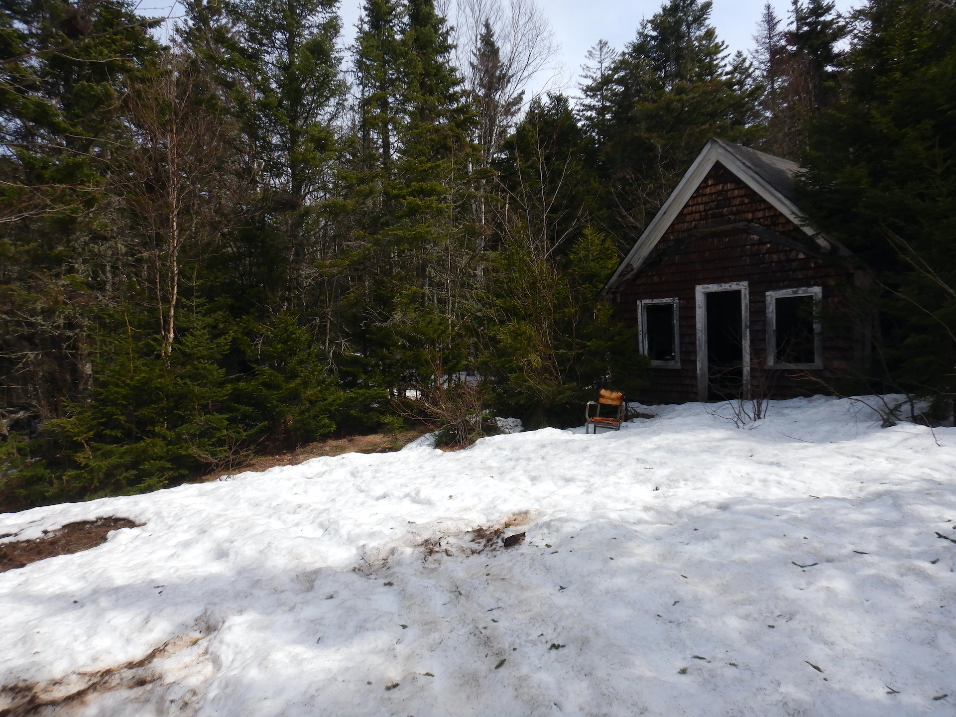 A derelict cabin in a forest. Wet, late season snow covered the bare area in front of the cabin. A mixed forest surround the cabin. The windows and door of the cabin is missing and the brick red lead paint is peeling from the outside.