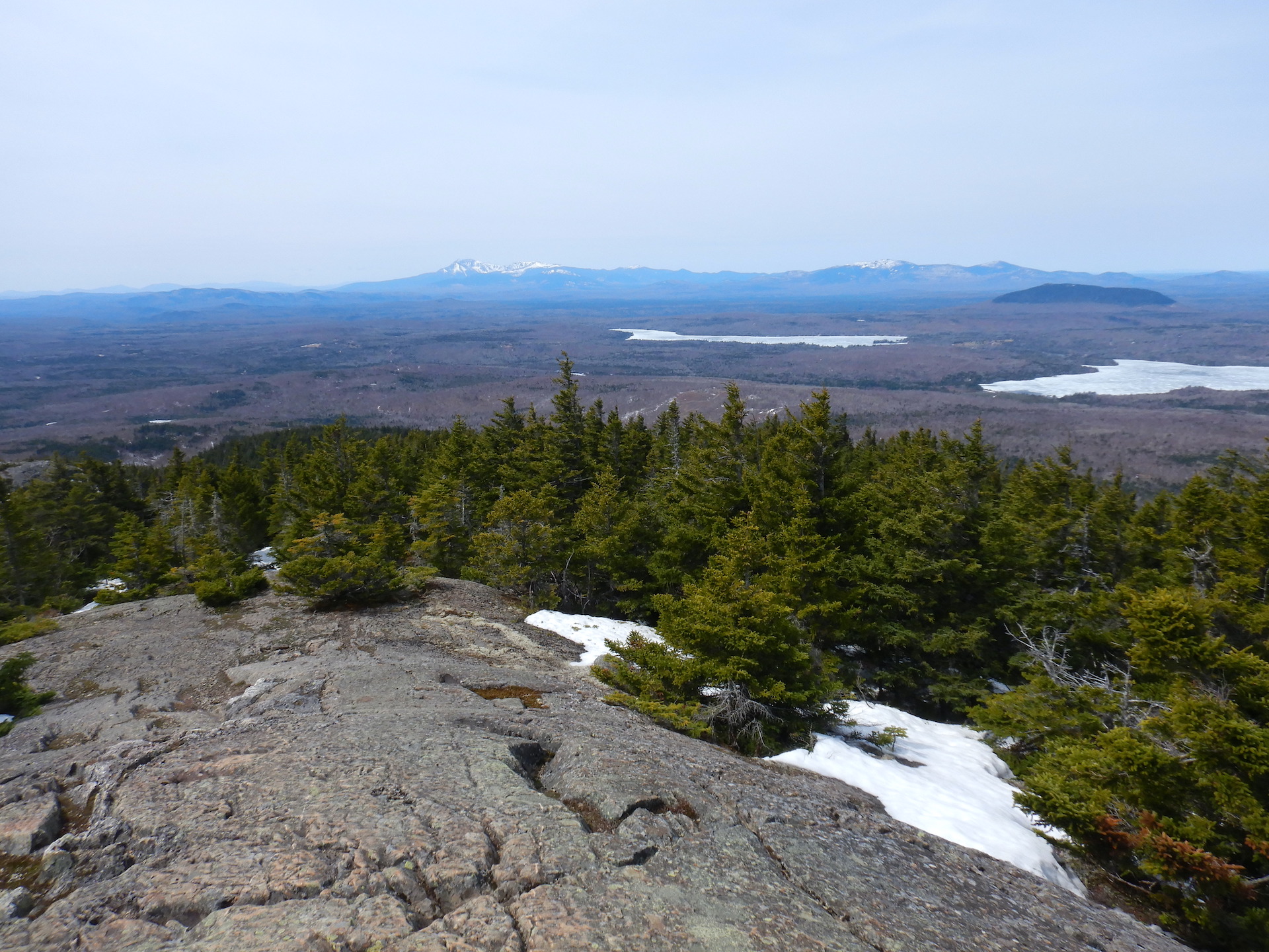 View from a mountain of a forested landscape. A ridge of large snow capped mountains form the horizon, although they appear small in the photo. Snow covered trees form the foreground just above windswept rock. Frozen ponds and forest fill the middle ground.