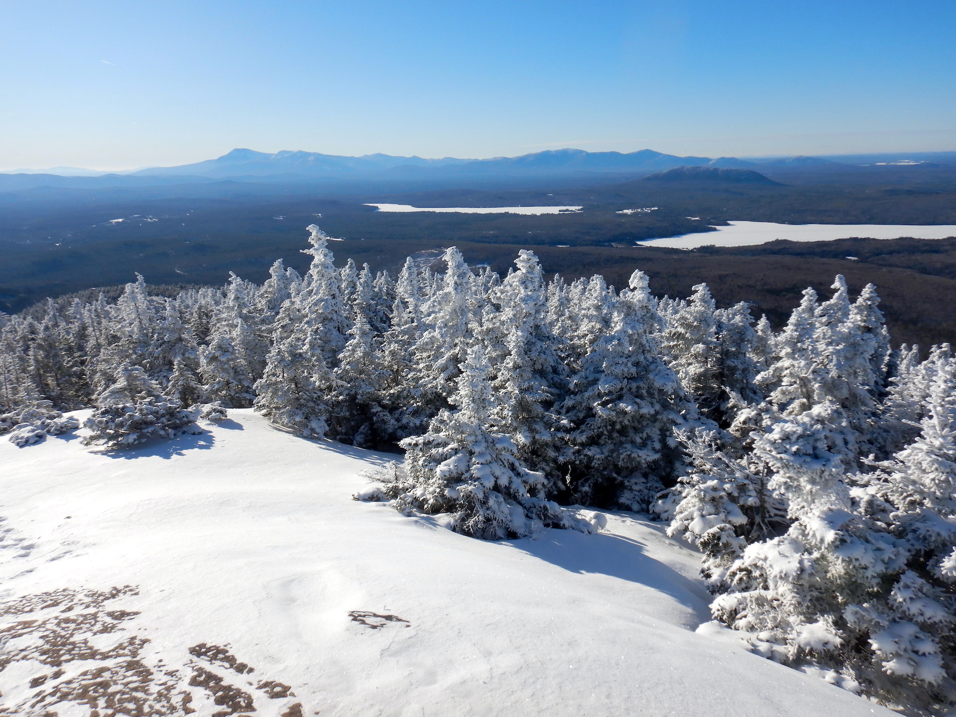 View from a mountain of a forested landscape. A ridge of large mountains form the horizon, although they appear small in the photo. Snow covered trees form the foreground just above windswept rock. Frozen ponds and forest sit in between the