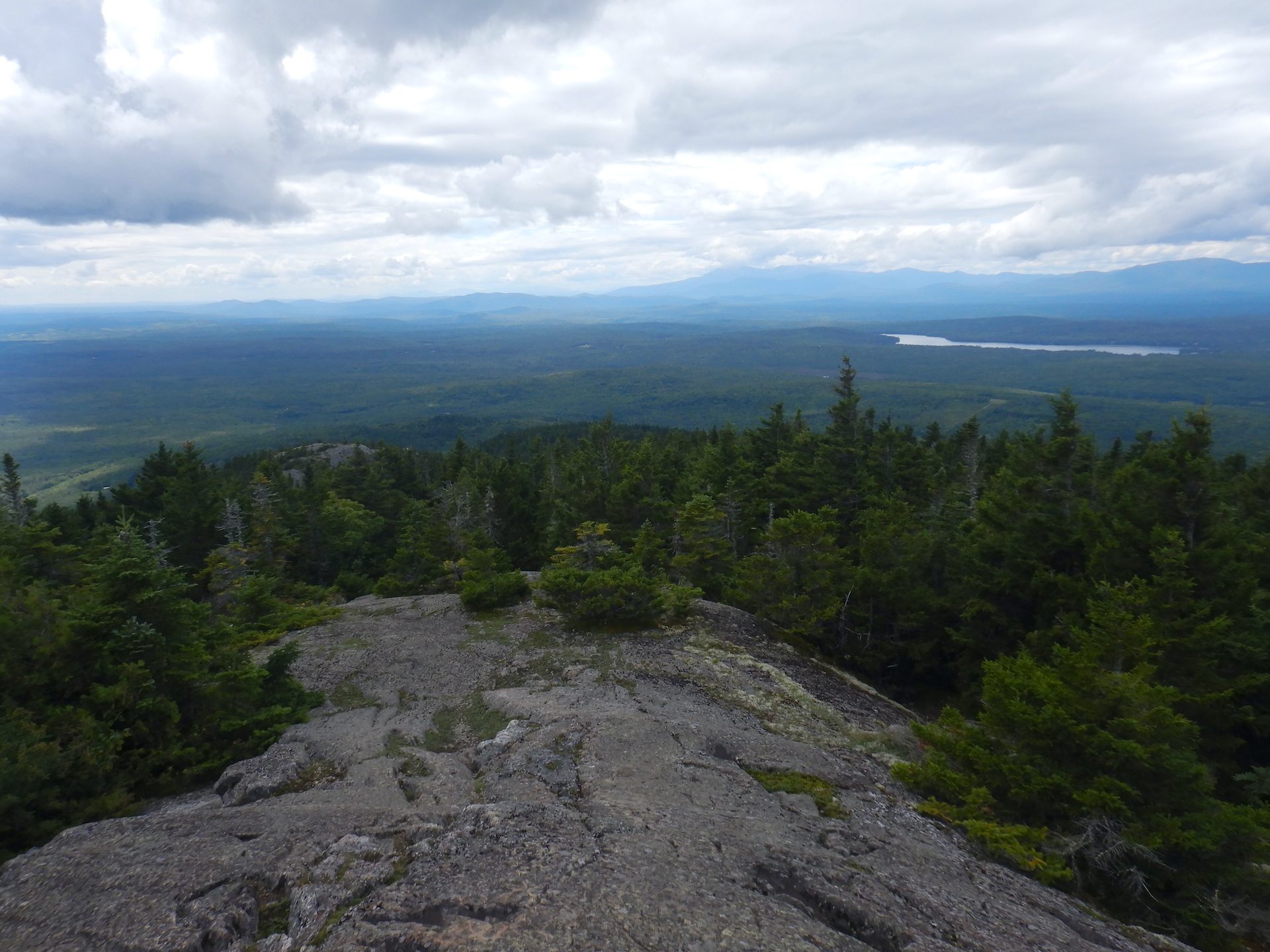 View of forested landscape from mountain summit. Bare rock covers the ground at bottom while spruce and fir trees cover the near slopes. The skies are mostly cloudy. Forest fills the lowlands. A pond and mountains can be seen near the horizon at center left.
