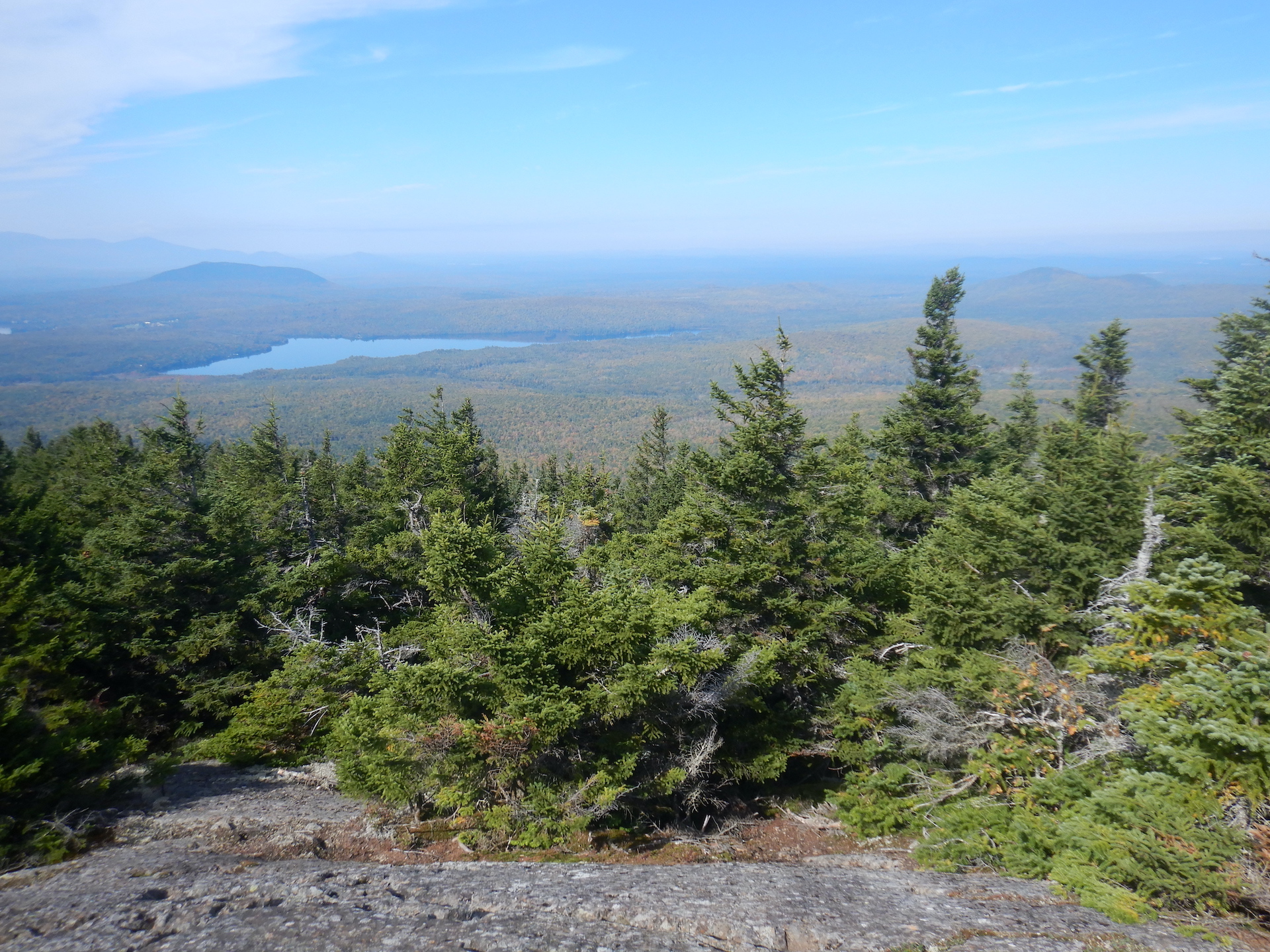 View of forested landscape from mountain summit. Bare rock covers the ground at bottom while spruce and fir trees cover the slopes. A pond is visible at center left. Haze obscures the horizon. The low elevation forest is speckled with yellow foliage.