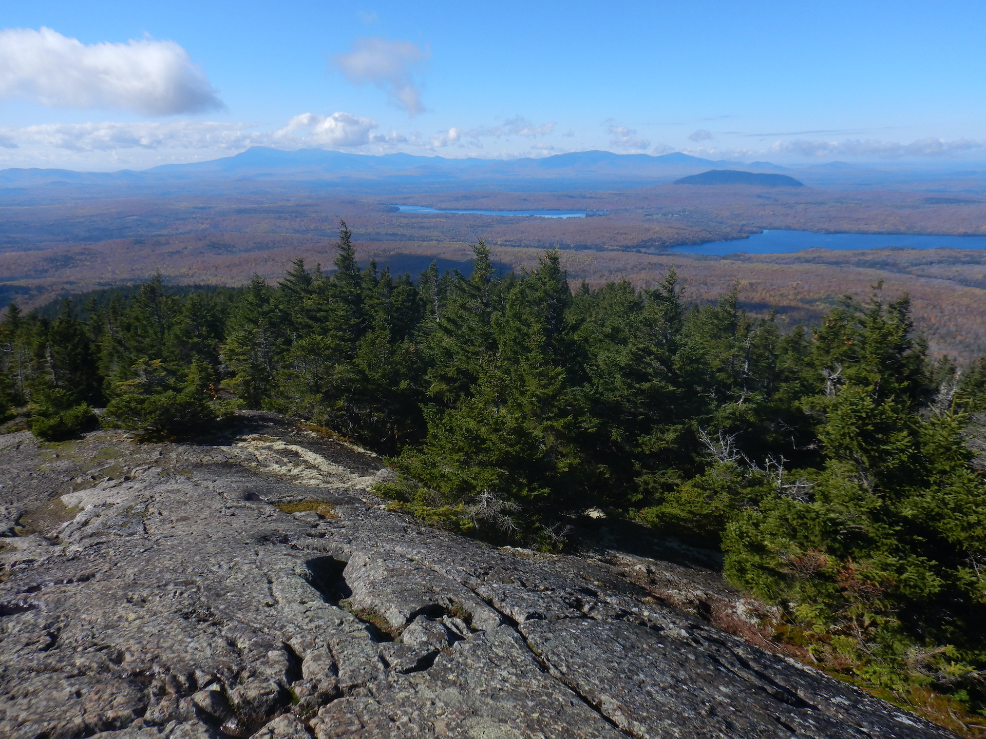 View of forested landscape from mountain summit. Bare rock covers the ground at bottom while spruce and fir trees cover the upper slopes. The lowland forest is mostly brown and bare of leaves. Tall mountains form the horizon although they look small due to the perspective. A pond is visible in the forest at center right.
