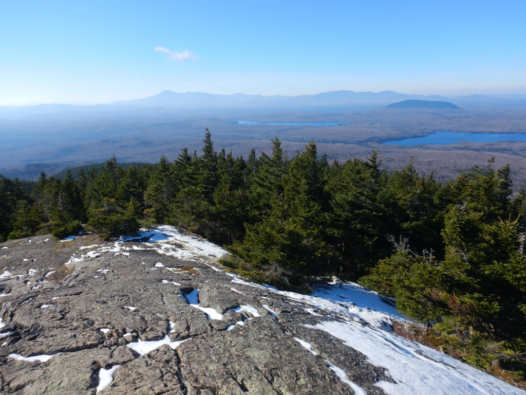 View of forested landscape from mountain summit. Bare rock with some patchy snow covers the ground at bottom while spruce and fir trees cover the upper slopes. The lowland forest is bare of leaves. Tall mountains form the horizon although they look small due to the perspective. A pond is visible in the forest at center right.
